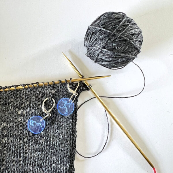 Knitting with grey yarn. There are two stitch markers with latch back hooks. Both markers are hooked round stitches.