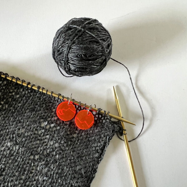 Knitting with grey yarn. Two orange perspex stitch makers are hand round the knitting pin.