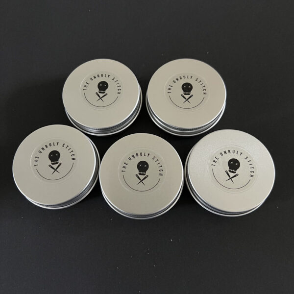 Five round screw top metal tins with The Unruly Stitch logo