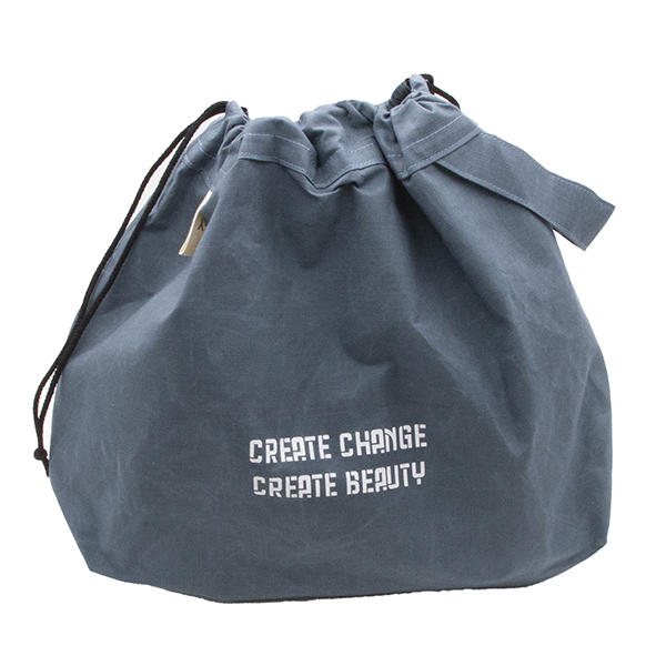 Bag with drawstrings closed. Large slate blue drawstring project bag. The bag is printed with text "CREATE CHANGE CREATE BEAUTY" in white ink. The bag has a handle, a drawstring and a cream label with The Unruly Stitch logo