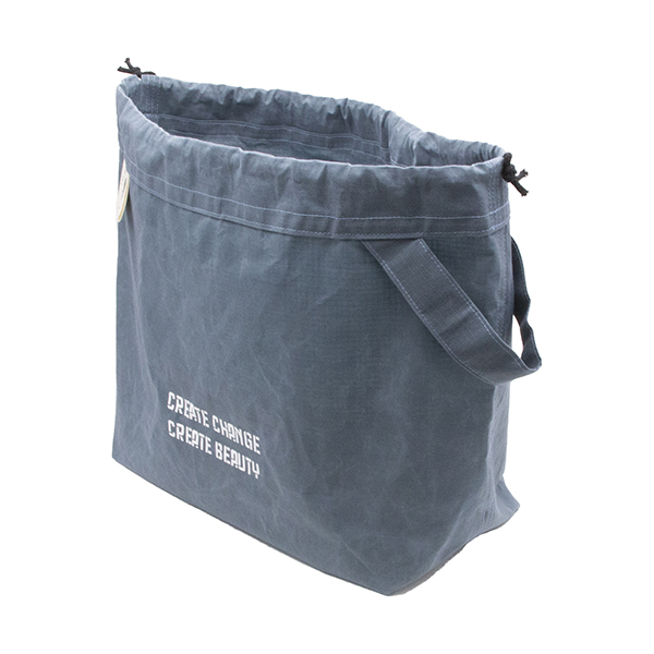 side view of large slate blue drawstring project bag. The bag is printed with text "CREATE CHANGE CREATE BEAUTY" in white ink. The bag has a handle, a drawstring and a cream label with The Unruly Stitch logo
