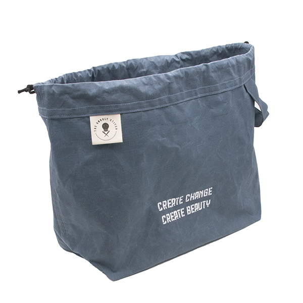large slate blue drawstring project bag. The bag is printed with text "CREATE CHANGE CREATE BEAUTY" in white ink. The bag has a handle, a drawstring and a cream label with The Unruly Stitch logo