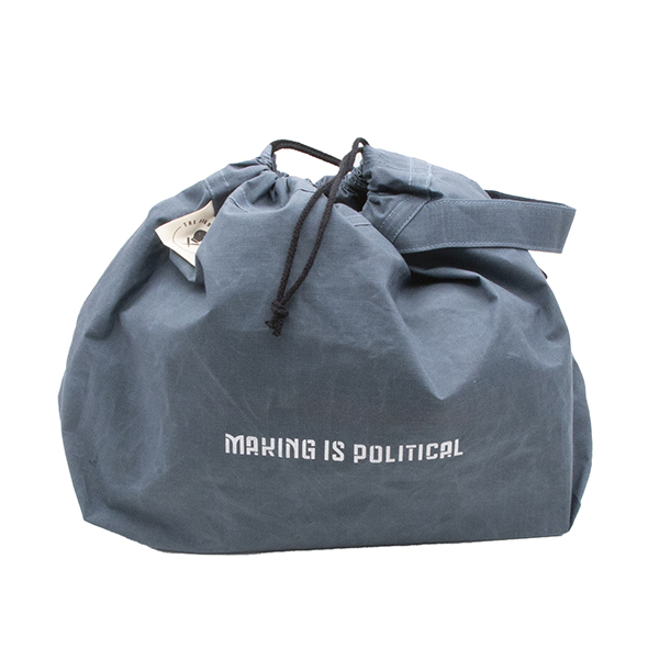 Bag with drawstrings closed. Large slate blue drawstring project bag. The bag is printed with text "MAKING IS POLITICAL" in white ink. The bag has a handle, a drawstring and a cream label with The Unruly Stitch logo