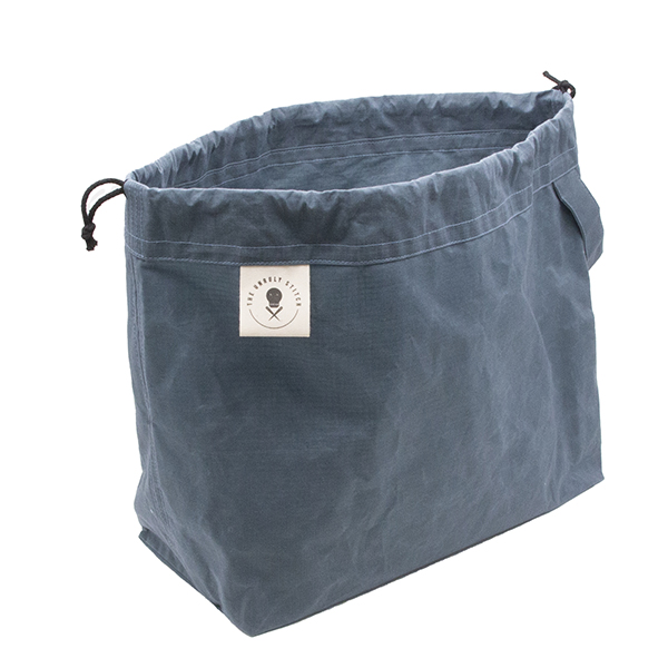 Front view of small slate blue drawstring project bag. The bag has a handle, a drawstring and a cream label with The Unruly Stitch logo