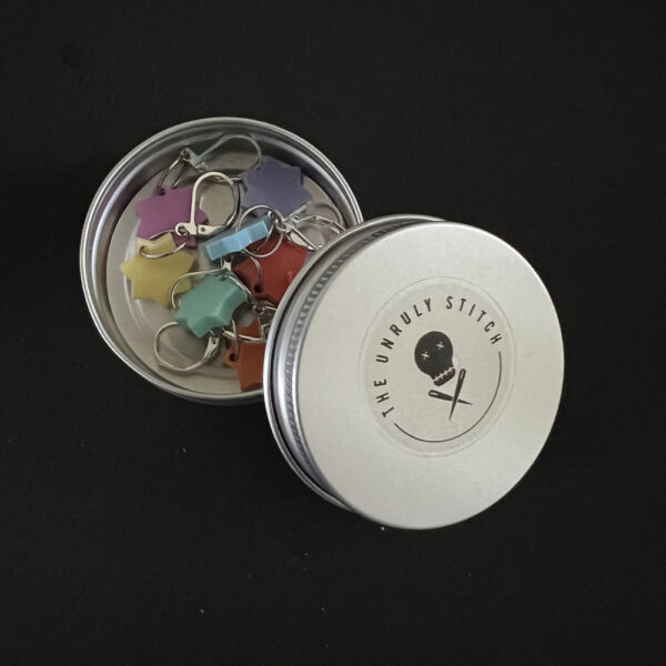 round screw top metal tins with The Unruly Stitch logo. The lid is open and sits to one side, showing the inside of the tin which contains stitch markers.