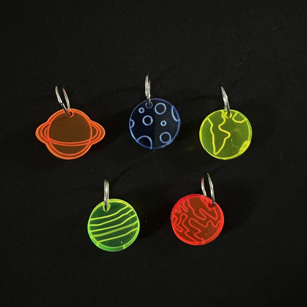 Five planets laser cut from fluorescent orange, blue, yellow, green and red perspex, . Mounted on f jump rings to create stitch markers.