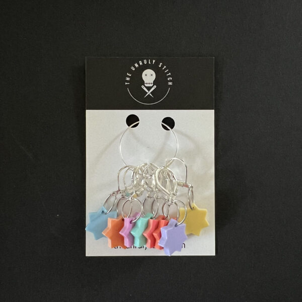 Eight stars laser cut from pastel perspex, Each star has seven points and is a different pastel colour. Mounted on latch back hooks to create stitch markers. Gathered together with a metal loop and displayed on a card with The Unruly Stitch logo