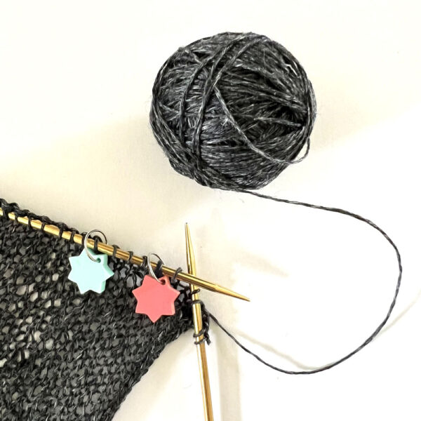 knitting with dark grey yarn showing two star stitch makers on jump rings looped over the needle.