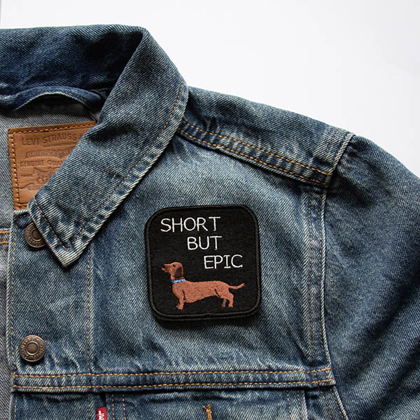 Short haired dachshund with the words short but epic embroidered patch on black felt on a denim jacket.