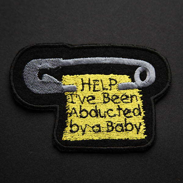 silver safety pin with scrap of yellow fabric with the words help ive been abducted by a baby embroidered patch on black felt on a black background.