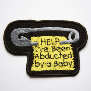 silver safety pin with scrap of yellow fabric with the words help ive been abducted by a baby embroidered patch on black felt.