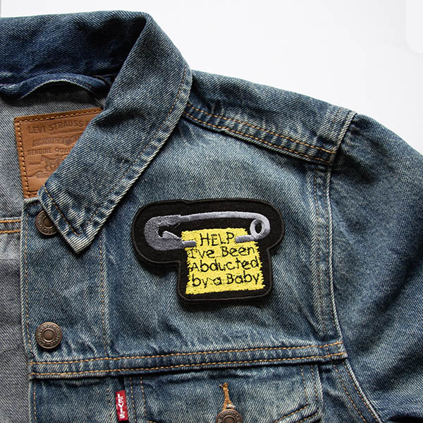 silver safety pin with scrap of yellow fabric with the words help ive been abducted by a baby embroidered patch on black felt on a denim jacket.