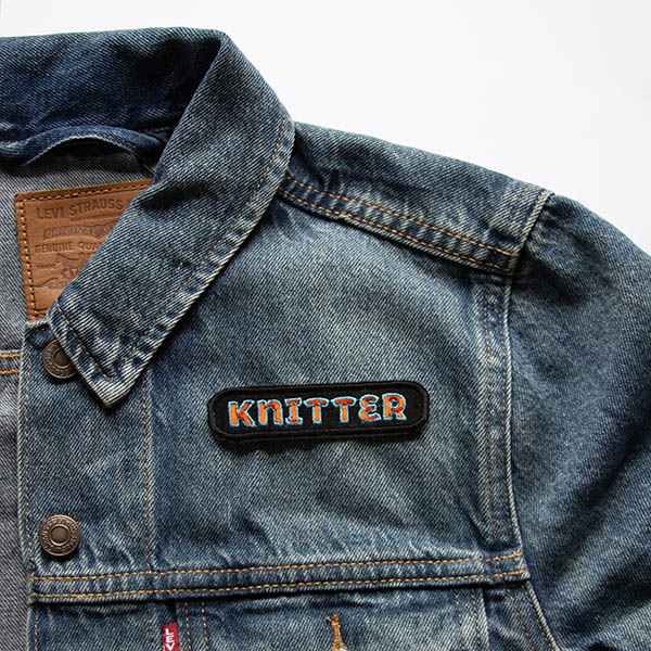 Knitter orange and turquoise embroidered patch on black felt on a denim jacket.