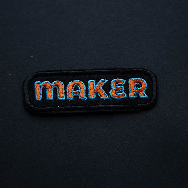 Maker embroidered patch in orange and turquoise on black felt on a black background.