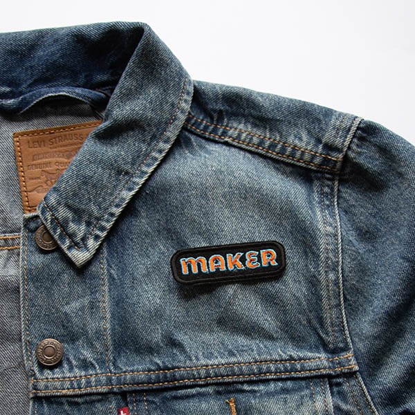 Maker embroidered patch in orange and turquoise on black felt on a denim jacket.