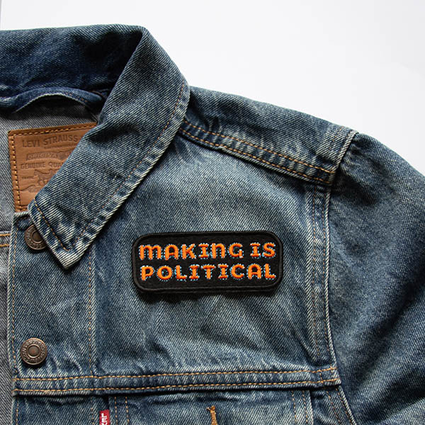 Making is political embroidered patch in orange and turquoise on black felt on a denim jacket.