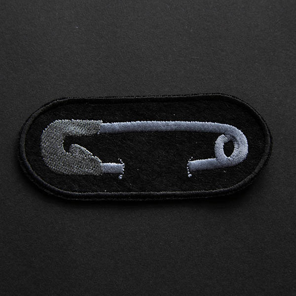 Embroidered patch showing silver safety pin on black felt on black background.