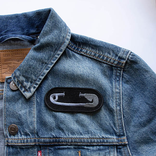 Embroidered patch showing silver safety pin on black felt attached to denim jacket.