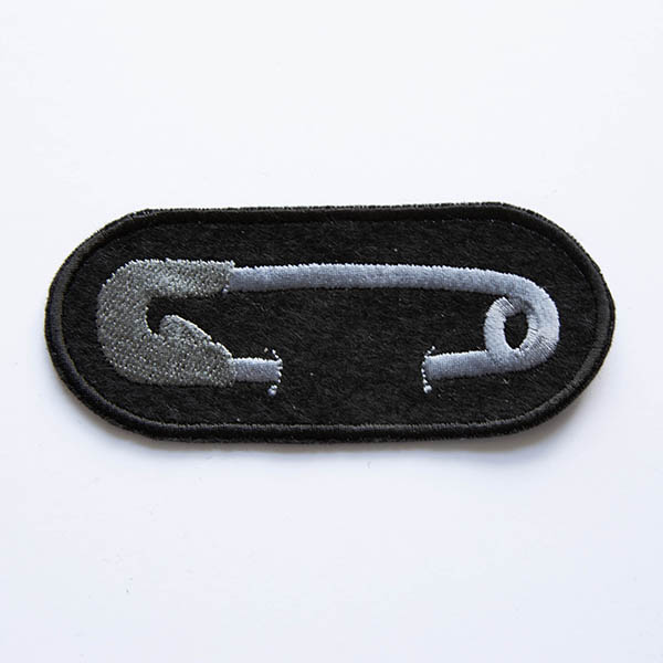 Embroidered patch showing silver safety pin on black felt.