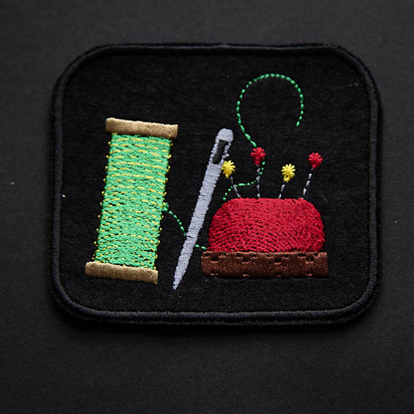 Thread needle and pin cushion embroidered on black felt patch on black background.