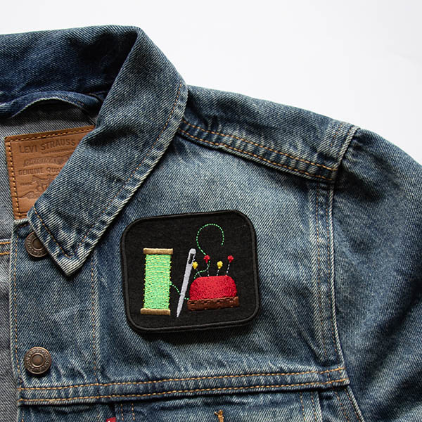 Thread needle and pin cushion embroidered on black felt patch on a denim jacket.