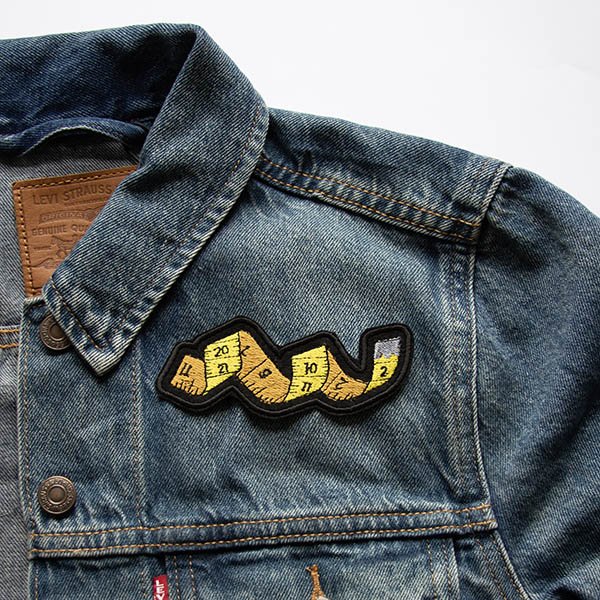 yellow tape measure embroidered patch with silver end and numbers on black felt on a denim jacket.