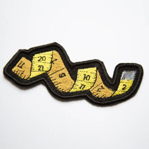 yellow tape measure embroidered patch with silver end and numbers on black felt.