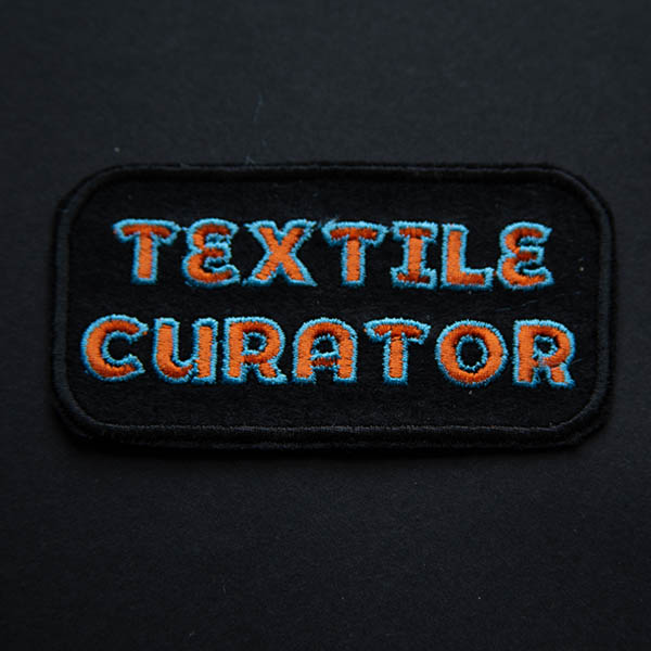 Textile curator embroidered in orange and turquoise patch on black felt and a black background.