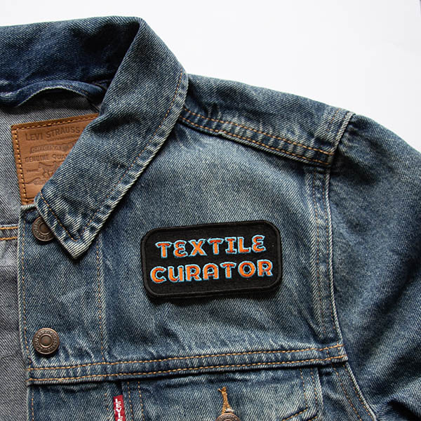 Textile curator embroidered in orange and turquoise patch on black felt background on a denim jacket.