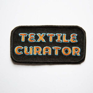 Textile curator embroidered in orange and turquoise patch on a black felt background.