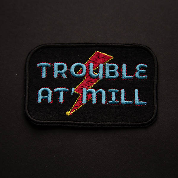 Trouble at mill overlaying lightening bolt embroidered patch on black felt on black background.