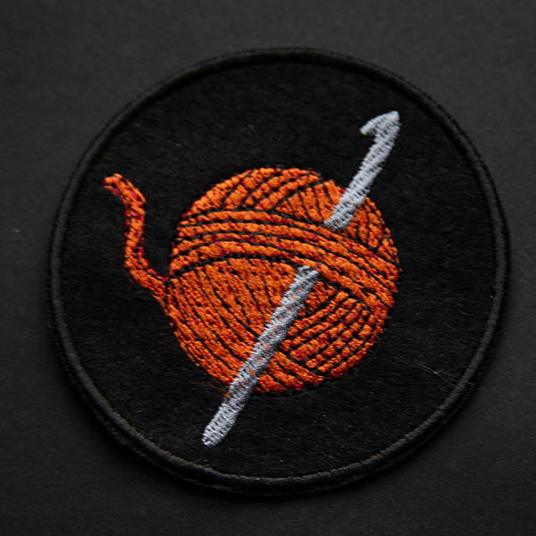 Embroidered patch showing an orange ball of yarn with a silver crochet hook on a black felt background shown on black background.