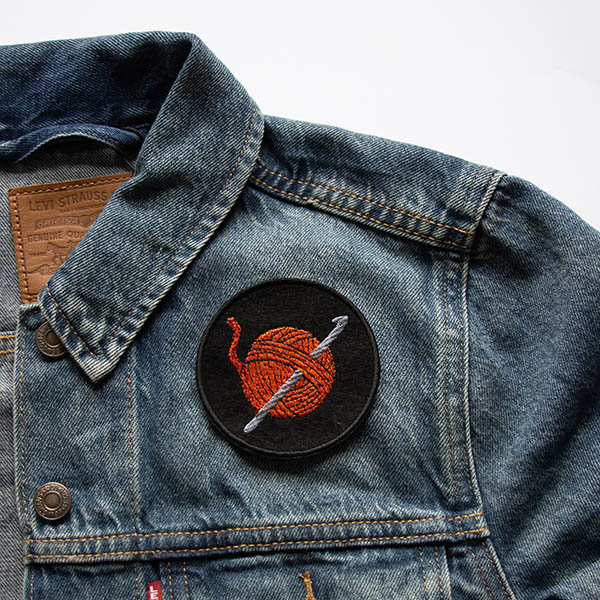 Embroidered patch showing an orange ball of yarn with a silver crochet hook on a black felt background on a denim jacket.