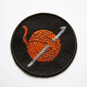 Embroidered patch showing an orange ball of yarn with a silver crochet hook on a black felt background.