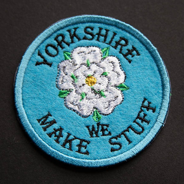 Embroidered patch showing a white rose with green leaves and yellow centre with yorkshire we make stuff on blue felt shown on black background.