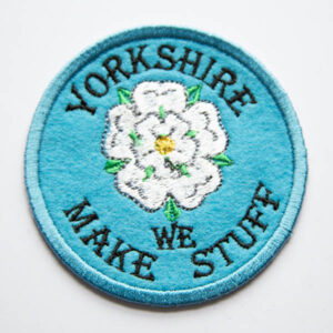 Embroidered patch showing a white rose with green leaves and yellow centre with yorkshire we make stuff on a blue felt background.