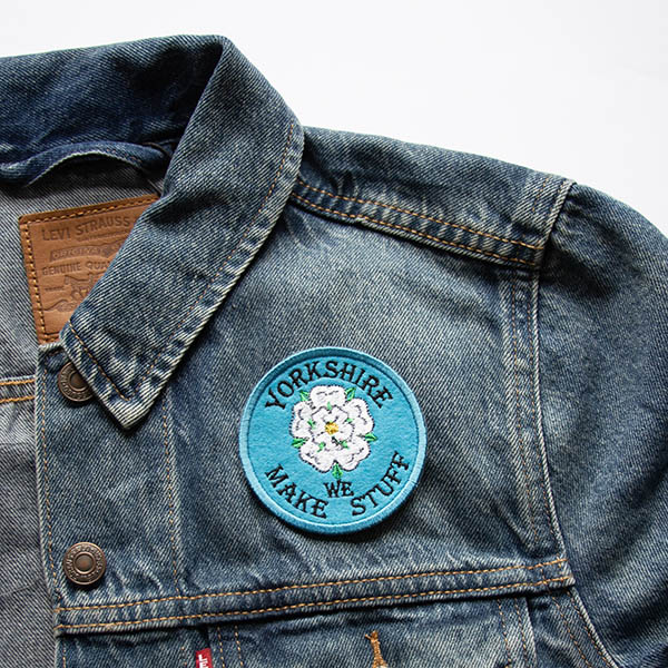 Embroidered patch showing a white rose with green leaves and yellow centre with yorkshire we make stuff on a blue felt background attached to a denim jacket.