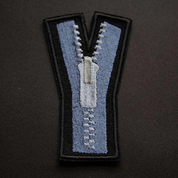 Embroidered patch showing a blue zip with silver teeth and pull on black felt on a black background.