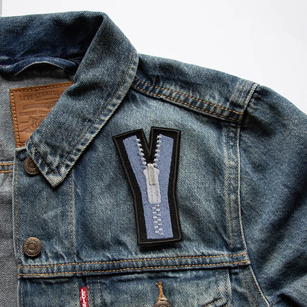 Embroidered patch showing a blue zip with silver teeth and pull on black felt attached to a denim jacket.