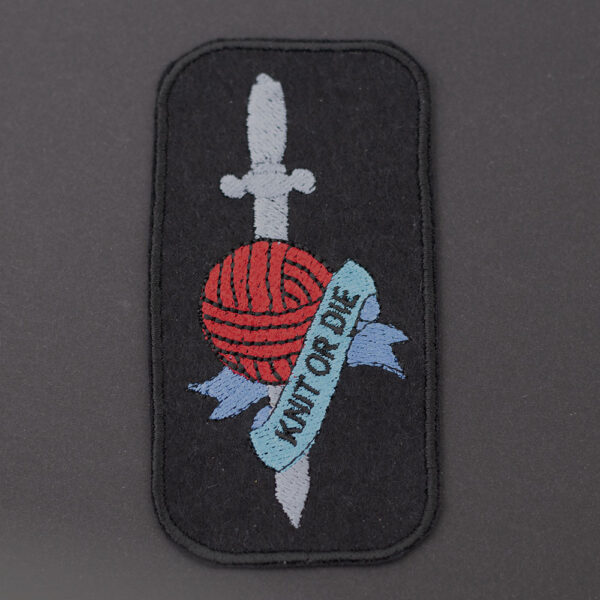 Embroidered patch on black felt showing tattoo style dagger and ball of yarn with banner. Text reads " KNIT OR DIE" . Shown on a dark background.