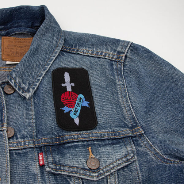 Embroidered patch on black felt showing tattoo style dagger and ball of yarn with banner. Text reads " KNIT OR DIE" . Attached to a denim jacket.