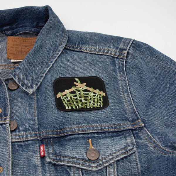Embroidered patch showing knitting on knitting pins on black felt attached to a denim jacket.