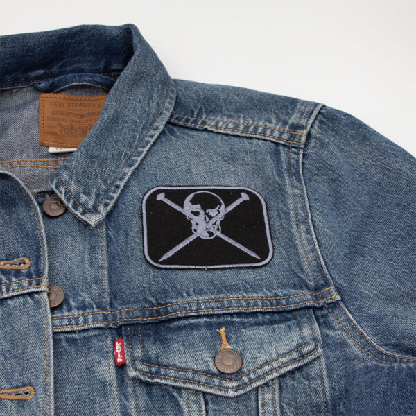 Embroidered patch showing a skull and crossed knitting pins on black felt attached to a denim jacket.