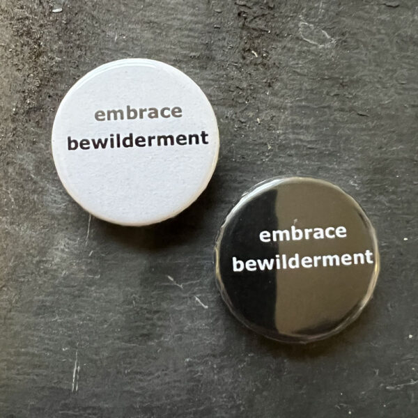 Two pin badges with the words embrace bewilderment. Black text, white background and white text, black background.