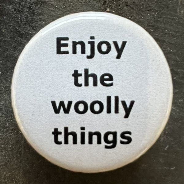Pin badge with the words Enjoy the woolly things. Black text, white background.