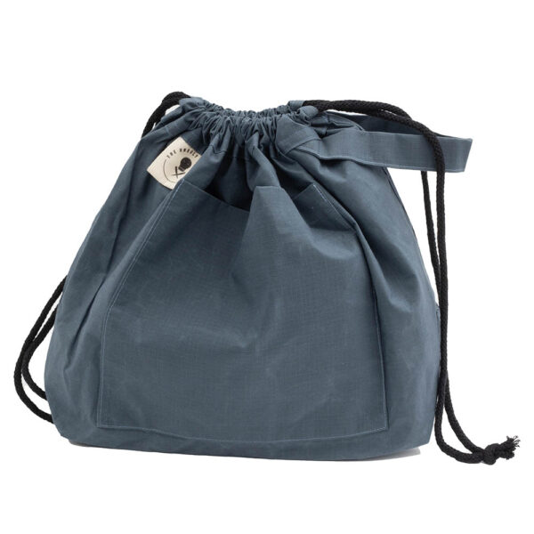 Bag with drawstring pulled closed. large slate blue drawstring project bag. The bag is has a large pocket on the front, a handle, a drawstring and a cream label with The Unruly Stitch logo