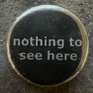 Pin badge with the words nothing to see here. Grey text, black background.