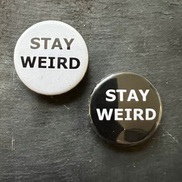Two pin badges with the words STAY WEIRD. Black text, white background and white text, black background.
