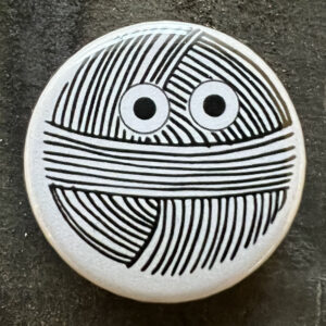 Pin badge with a line drawing of a ball of yarn with googly eyes. Black lines, white background.
